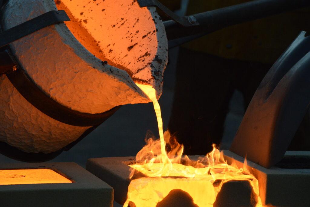 Ein Schmelzofen wird geöffnet und das glühend heiße Metall wird in eine Form gegossen, während Flammen aufsteigen.